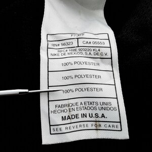 Pu&ograve; includere: Primo piano di un'etichetta di abbigliamento bianca con testo nero. L'etichetta include il testo "RN# 56323", "CA# 05553", "100% POLYESTER" e "MADE IN U.S.A.". Uno strumento di metallo punta all'etichetta.