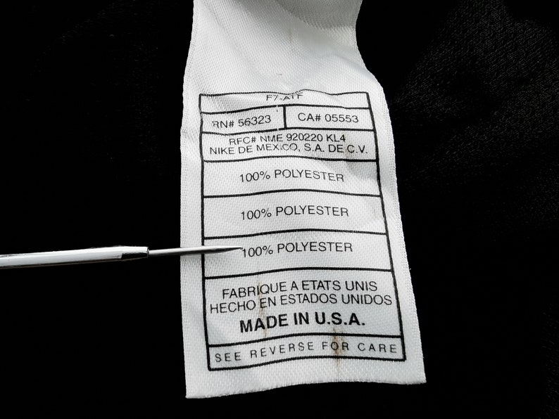 Pu&ograve; includere: Primo piano di un'etichetta di abbigliamento bianca con testo nero. L'etichetta include il testo "RN# 56323", "CA# 05553", "100% POLYESTER" e "MADE IN U.S.A.". Uno strumento di metallo punta all'etichetta.