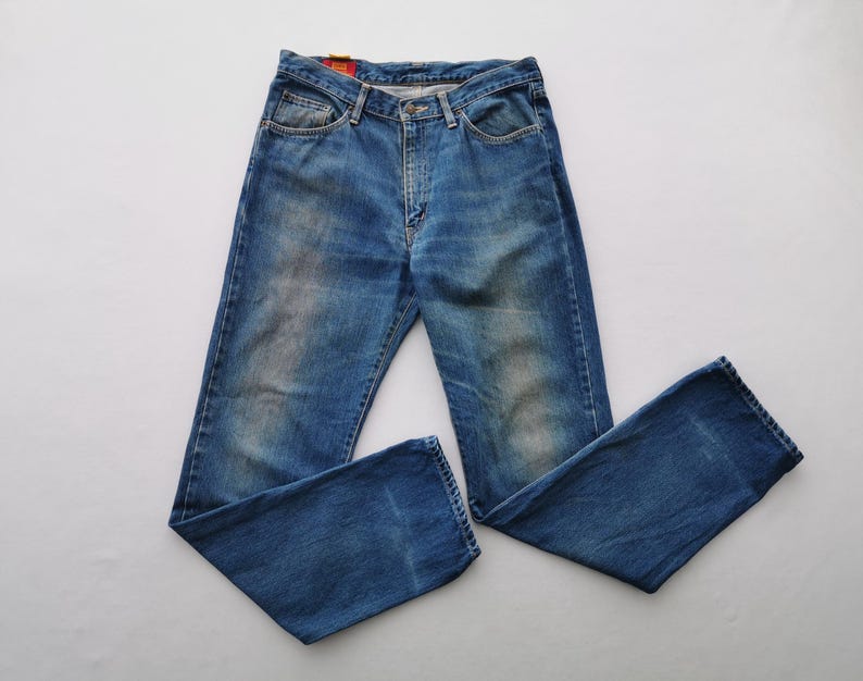 Puede incluir: Vaqueros azules de mezclilla con un dise&ntilde;o cl&aacute;sico de cinco bolsillos. Los vaqueros tienen un lavado descolorido y un corte de pierna recta. Los vaqueros est&aacute;n planos sobre una superficie blanca. Cierre de bot&oacute;n y cremallera.