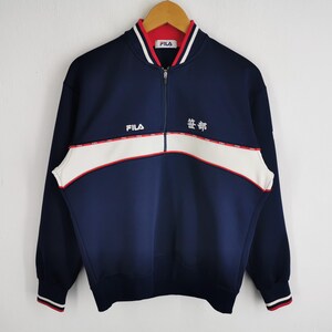 May include: Navy blue zip-up sweatshirt with white and red accents. Features the brand name "FILA" and Japanese text. The sweatshirt has a white stripe across the chest and red and white striped cuffs.