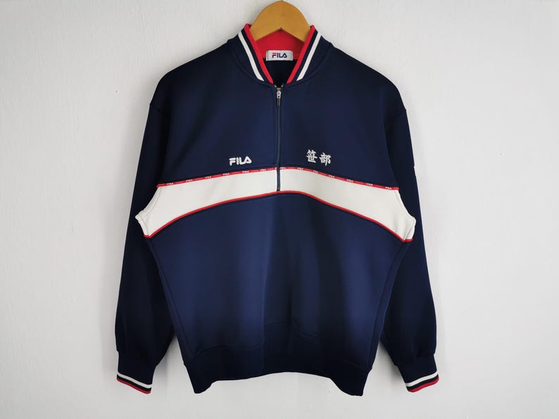 May include: Navy blue zip-up sweatshirt with white and red accents. Features the brand name "FILA" and Japanese text. The sweatshirt has a white stripe across the chest and red and white striped cuffs.