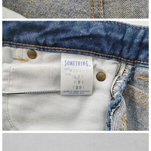 May include: Close-up of denim jeans with multiple tags. The top tag reads "TURQUOISE BLUE SOMETHING PRODUCED BY EDWIN." Other tags provide size information, including a waist size of W63, and care instructions. The jeans are made of 100% cotton.