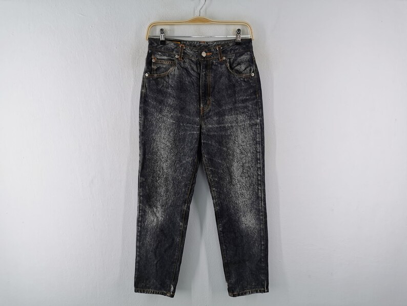 May include: Acid wash denim jeans with a high waist and straight leg. The jeans are a dark gray color with lighter gray, mottled patterns throughout. They are hanging on a wooden hanger against a white background.