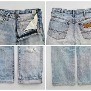 May include: Light blue denim Wrangler jeans. The jeans have a faded wash, a zipper fly, and a distressed area on the right leg. The back features two pockets and a Wrangler patch.