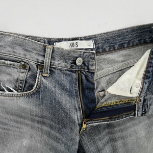 May include: Close-up of a pair of faded gray denim jeans with a partially unzipped fly. The jeans have a classic five-pocket design, a button closure, and a small label that reads "XU-S". The denim has a worn, distressed look.
