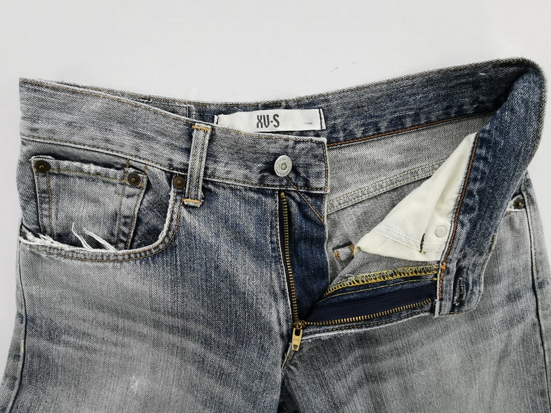 May include: Close-up of a pair of faded gray denim jeans with a partially unzipped fly. The jeans have a classic five-pocket design, a button closure, and a small label that reads "XU-S". The denim has a worn, distressed look.
