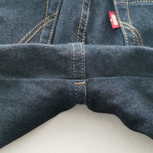 K&ouml;nnte beinhalten: Nahaufnahme einer dunkelblauen Jeans. Die Jeans hat beige N&auml;hte und ein rotes Etikett. Die N&auml;hte sind an den N&auml;hten und Taschen sichtbar. Der Denim hat ein leicht strukturiertes Aussehen.