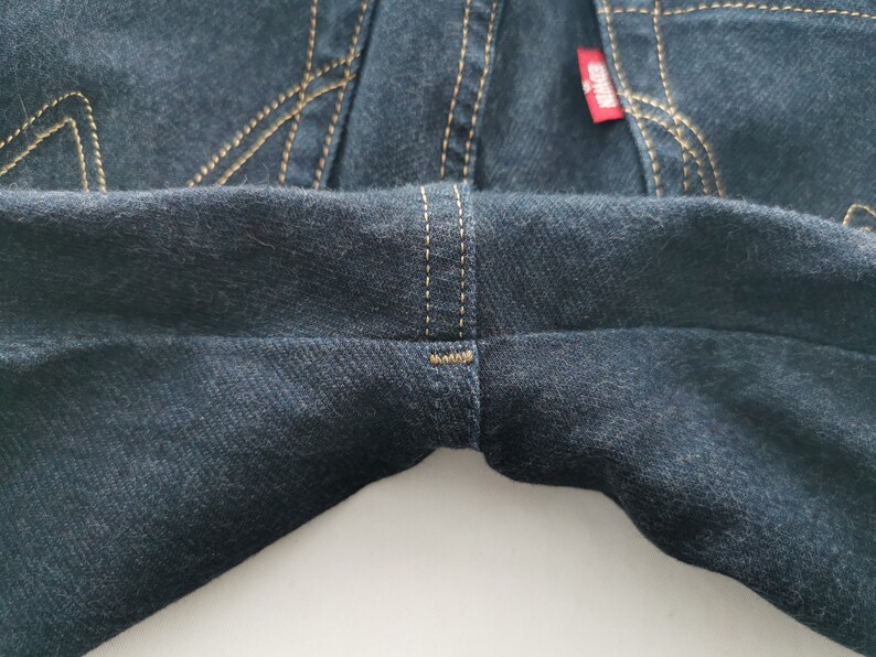 K&ouml;nnte beinhalten: Nahaufnahme einer dunkelblauen Jeans. Die Jeans hat beige N&auml;hte und ein rotes Etikett. Die N&auml;hte sind an den N&auml;hten und Taschen sichtbar. Der Denim hat ein leicht strukturiertes Aussehen.