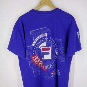 May include: A royal blue t-shirt with a graphic design on the back. The design includes the FILA logo, the text "TAKE NO PRISONERS", and other text. The shirt is hanging on a wooden hanger.