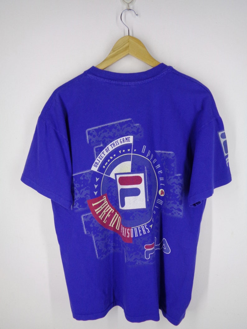 May include: A royal blue t-shirt with a graphic design on the back. The design includes the FILA logo, the text "TAKE NO PRISONERS", and other text. The shirt is hanging on a wooden hanger.