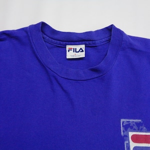 May include: A royal blue t-shirt with a crew neck collar. The shirt features the FILA logo in white, red, and blue on the lower right. The FILA brand label is visible on the inside collar.
