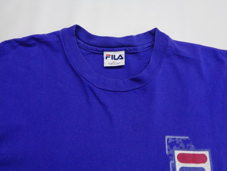 May include: A royal blue t-shirt with a crew neck collar. The shirt features the FILA logo in white, red, and blue on the lower right. The FILA brand label is visible on the inside collar.