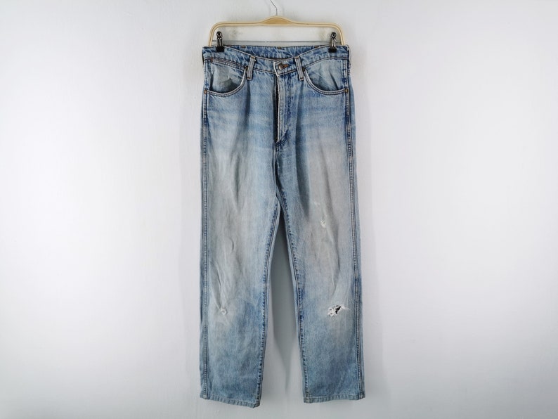 May include: Light blue denim jeans with a faded wash and distressed details, including rips and wear marks. The jeans are hanging on a wooden hanger against a white background. Classic five-pocket design.