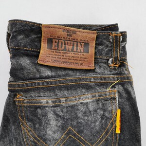 May include: Close-up of a pair of black denim jeans with a faded wash effect. The jeans feature orange stitching and a brown leather patch with the brand name "EDWIN" and text. A yellow tag is visible on the back pocket.