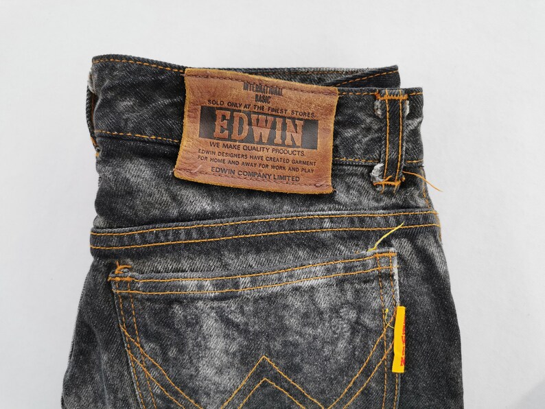 May include: Close-up of a pair of black denim jeans with a faded wash effect. The jeans feature orange stitching and a brown leather patch with the brand name "EDWIN" and text. A yellow tag is visible on the back pocket.