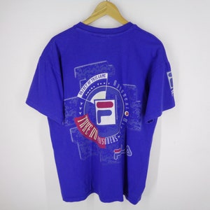 May include: A royal blue t-shirt with a graphic design featuring the FILA logo in white, red, and blue. The design includes the text "OBJECT OF THIS GAME" and "TAKE IT TO THE PRISONERS". The shirt is hanging on a wooden hanger.
