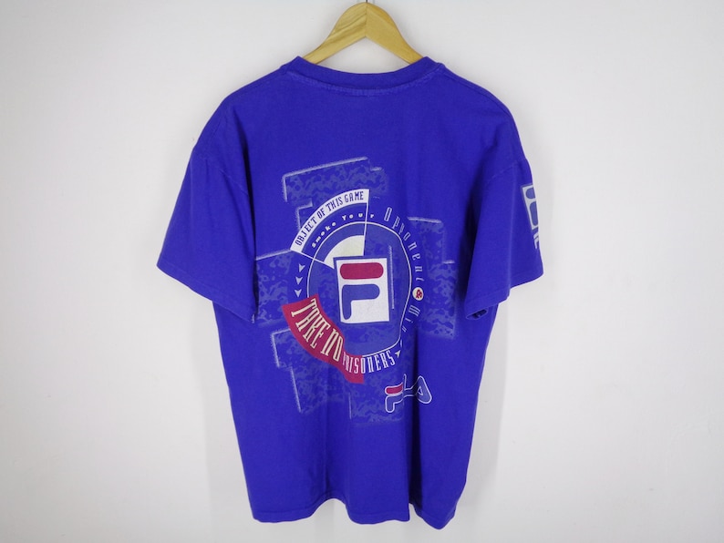 May include: A royal blue t-shirt with a graphic design featuring the FILA logo in white, red, and blue. The design includes the text "OBJECT OF THIS GAME" and "TAKE IT TO THE PRISONERS". The shirt is hanging on a wooden hanger.