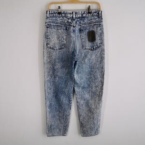 May include: Acid wash denim jeans hanging on a wooden hanger. The jeans have a light blue and white marbled pattern. The back pockets and a small patch are visible. The jeans are a vintage style.