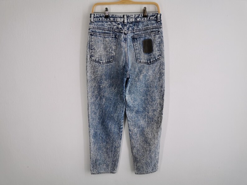 May include: Acid wash denim jeans hanging on a wooden hanger. The jeans have a light blue and white marbled pattern. The back pockets and a small patch are visible. The jeans are a vintage style.