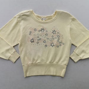 May include: Pale yellow sweatshirt with long sleeves and a crew neck. The front features a floral design with the word "courreges" in the center. The cuffs and hem are ribbed.