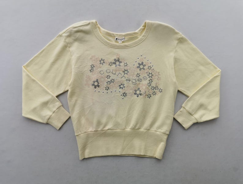 May include: Pale yellow sweatshirt with long sleeves and a crew neck. The front features a floral design with the word "courreges" in the center. The cuffs and hem are ribbed.
