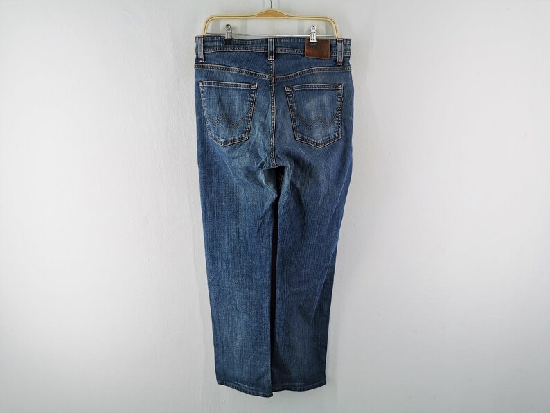 May include: Blue denim jeans hanging on a wooden hanger. The jeans have a classic five-pocket design, a straight leg, and a brown leather patch on the back. The jeans are a medium wash and appear to be unworn.