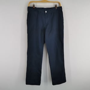 May include: Navy blue pants hanging on a wooden hanger against a white wall. The pants have a button and zipper closure, with pockets on the front and back. The fabric appears to be a sturdy cotton or blend.