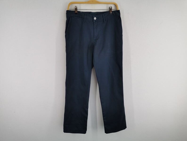 May include: Navy blue pants hanging on a wooden hanger against a white wall. The pants have a button and zipper closure, with pockets on the front and back. The fabric appears to be a sturdy cotton or blend.