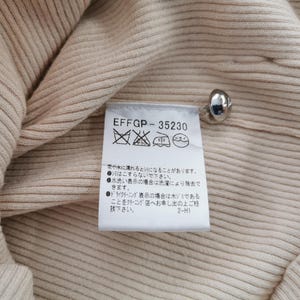 May include: Close-up of a beige ribbed knit fabric with a silver button and a care label. The label has the text "EFFGP-35230" and washing instructions in Japanese.