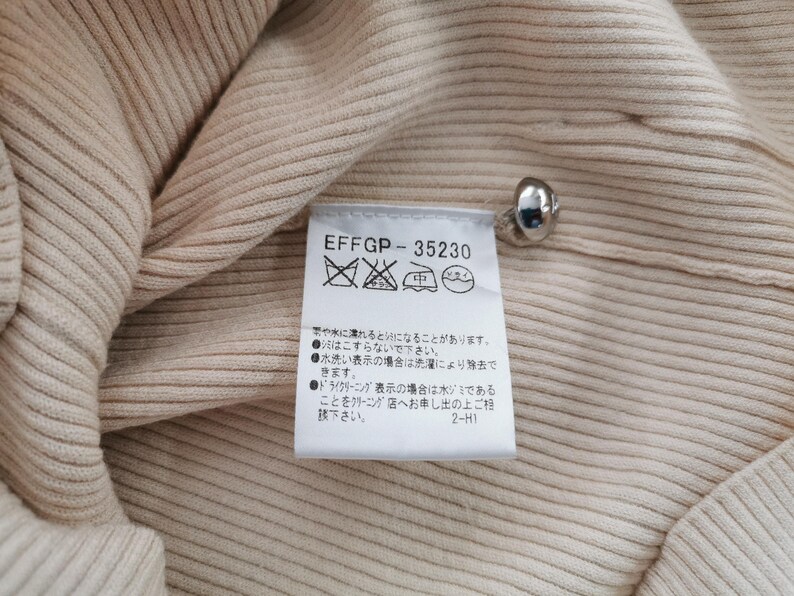 May include: Close-up of a beige ribbed knit fabric with a silver button and a care label. The label has the text "EFFGP-35230" and washing instructions in Japanese.
