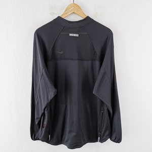 Pu&ograve; includere: Maglia sportiva a maniche lunghe grigio scuro con un piccolo logo Nike. La maglia ha un pannello in rete sulla parte superiore della schiena e una piccola etichetta. Presenta due tasche laterali con cerniere. La maglia &egrave; appesa a una gruccia di legno.