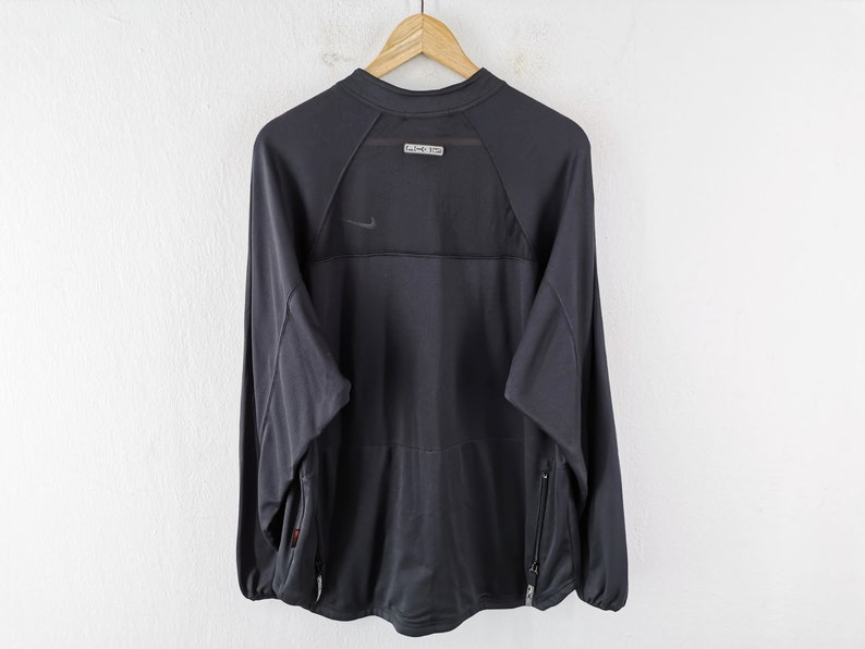 Pu&ograve; includere: Maglia sportiva a maniche lunghe grigio scuro con un piccolo logo Nike. La maglia ha un pannello in rete sulla parte superiore della schiena e una piccola etichetta. Presenta due tasche laterali con cerniere. La maglia &egrave; appesa a una gruccia di legno.