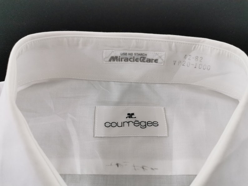 May include: Close-up of a white dress shirt collar with the brand label "courr&egrave;ges" visible. The shirt has a "Miracle Care" label and size markings "42-82 VP20-1000". The shirt is made of a smooth, likely cotton, fabric.