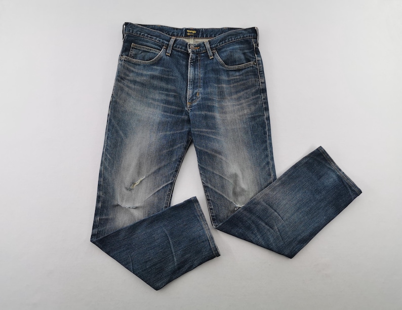May include: Blue denim jeans with a faded wash and distressed details. The jeans have a classic five-pocket design and a button and zipper closure. The jeans are laid flat on a white surface.