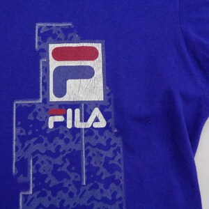 May include: A royal blue t-shirt featuring the FILA logo in white, red, and blue. The logo is slightly distressed, with a faded appearance. The shirt has short sleeves and a crew neck.