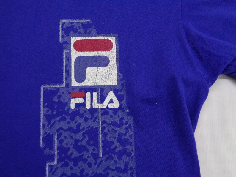 May include: A royal blue t-shirt featuring the FILA logo in white, red, and blue. The logo is slightly distressed, with a faded appearance. The shirt has short sleeves and a crew neck.