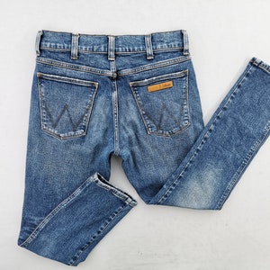 May include: Blue denim jeans with a classic five-pocket design. The jeans feature a straight leg cut, a button and zipper closure, and the Wrangler logo on the back pocket. The jeans are made of a durable denim material.