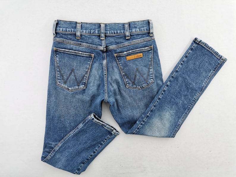May include: Blue denim jeans with a classic five-pocket design. The jeans feature a straight leg cut, a button and zipper closure, and the Wrangler logo on the back pocket. The jeans are made of a durable denim material.