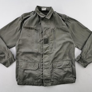 May include: An olive green jacket with a collar, buttoned cuffs, and several pockets. The jacket has two zipped chest pockets and two lower flap pockets. The fabric has a worn appearance, possibly vintage or military-inspired.