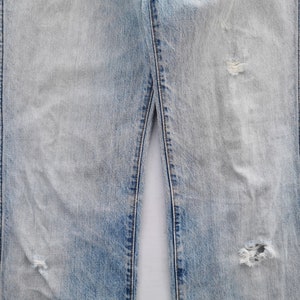 May include: Light blue denim jeans with distressed details, including rips and fading. The jeans have a classic five-pocket design and a straight leg fit. The fabric shows signs of wear.