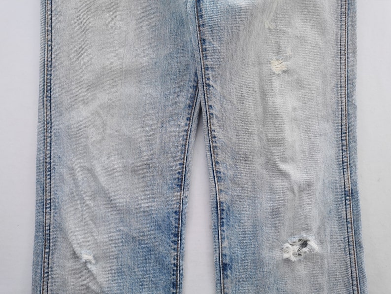 May include: Light blue denim jeans with distressed details, including rips and fading. The jeans have a classic five-pocket design and a straight leg fit. The fabric shows signs of wear.