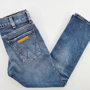 May include: Blue denim jeans with a classic five-pocket design. Features a brown leather patch with the "Wrangler" logo on the back pocket. The jeans have a straight leg cut and are made of a medium-wash denim.