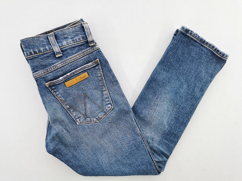 May include: Blue denim jeans with a classic five-pocket design. Features a brown leather patch with the "Wrangler" logo on the back pocket. The jeans have a straight leg cut and are made of a medium-wash denim.