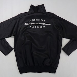 Puede incluir: Chaqueta bomber negra con cuello alto y cremallera completa. La parte trasera de la chaqueta presenta el texto blanco "A MOVIE FOR Cobraa-Lax ALL GENDERS". La chaqueta tiene mangas largas y dobladillo y puños acanalados.