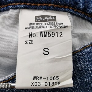May include: Close-up of a blue denim jeans label. The label reads "Wrangler" and includes the style number WM5912 and size "S". The label also includes the text "MADE UNDER LICENSE FROM WRANGLER APPAREL CORPORATION".