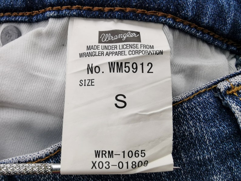 May include: Close-up of a blue denim jeans label. The label reads "Wrangler" and includes the style number WM5912 and size "S". The label also includes the text "MADE UNDER LICENSE FROM WRANGLER APPAREL CORPORATION".