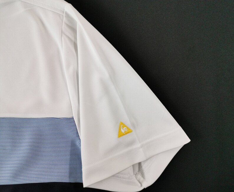 May include: White and blue short-sleeve polo shirt. The shirt features a white upper section, a blue striped middle section, and a black lower section. A yellow triangle logo with a rooster is on the sleeve.