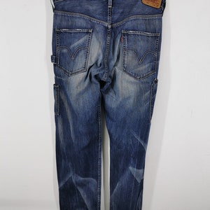 K&ouml;nnte beinhalten: Blaue Jeans aus Denim mit verwaschener Optik, mit zwei Ges&auml;&szlig;taschen mit dem Levi's-Logo und einem kleinen roten Etikett. Die Jeans haben eine Zimmermannstasche an der Seite und h&auml;ngen an einem Holzb&uuml;gel.