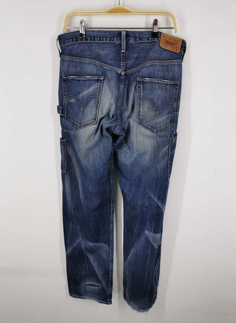 K&ouml;nnte beinhalten: Blaue Jeans aus Denim mit verwaschener Optik, mit zwei Ges&auml;&szlig;taschen mit dem Levi's-Logo und einem kleinen roten Etikett. Die Jeans haben eine Zimmermannstasche an der Seite und h&auml;ngen an einem Holzb&uuml;gel.