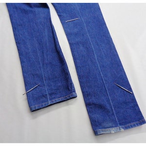 May include: Close-up shots of blue denim jeans. The jeans are laid out flat, showcasing the seams and the hem. The fabric appears to be a medium wash denim. Pins are used to hold the fabric together.